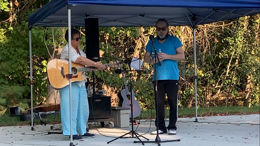 A woman and a man are standing under the tent getting ready to sing. The woman is adjusting an acoustic guitar.