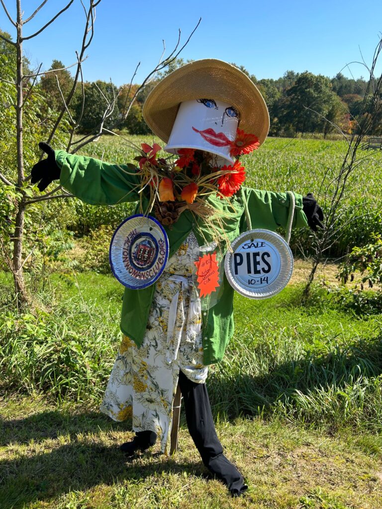 A feminine scarecrow with big red lipstick lips and a straw hat wears a green coat and print dress with pie plates hanging from each shoulder.