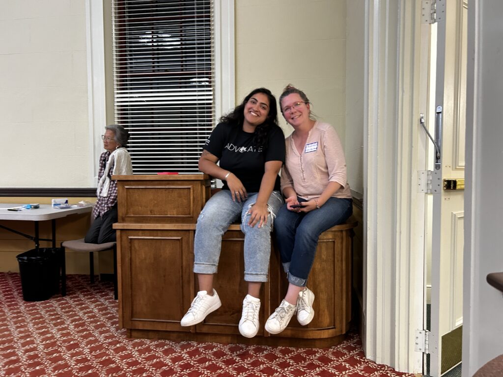 Two smiling women sitting on a desk at the entrance to Room 204. In the background, a third woman is perch