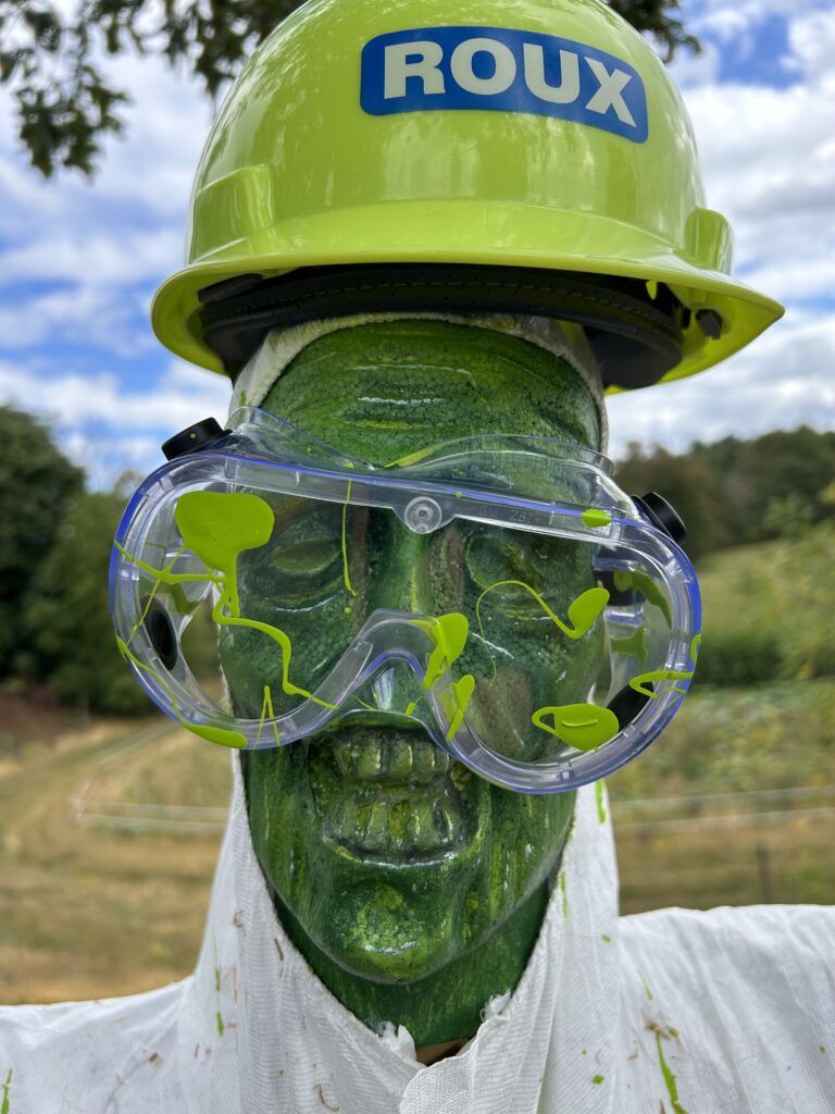 The Hazardous Waste Monster has a shiny green skull for a head and is wearing goggles splashed with green paint and a bright green Roux Associates hard hat. 