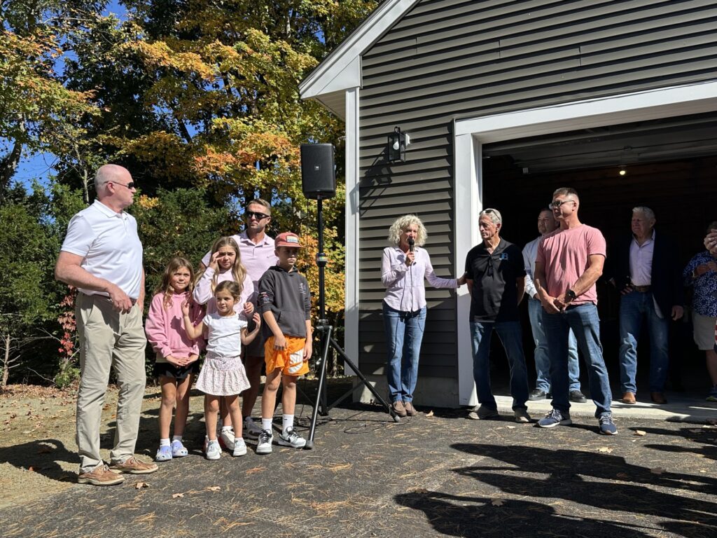 A woman wearing a pink button-down shirt and heans speaks in front of the garage of the newly built house. A number of people are listening to her speak.