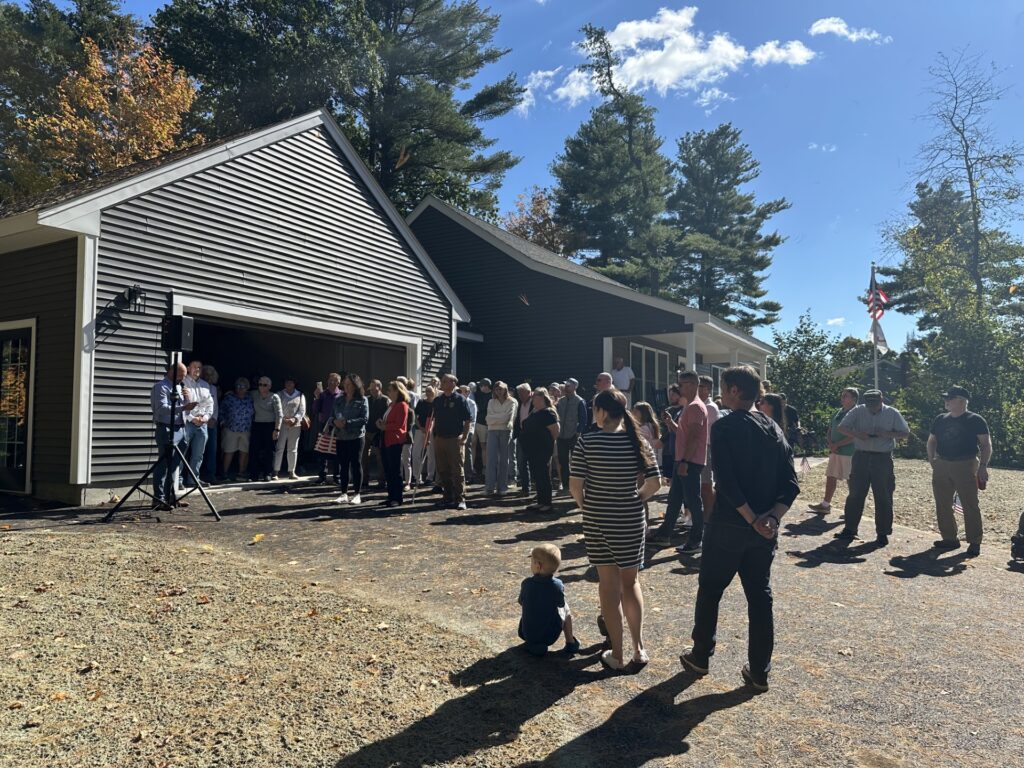 A group of people stand on the driveway of the new gray house. AB Veteran Services Officer James MacRae is speaking.