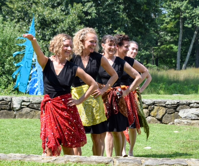 Five women wearing brightly colored scarves tied around their waists are dancing in a line outdoors in the sun.