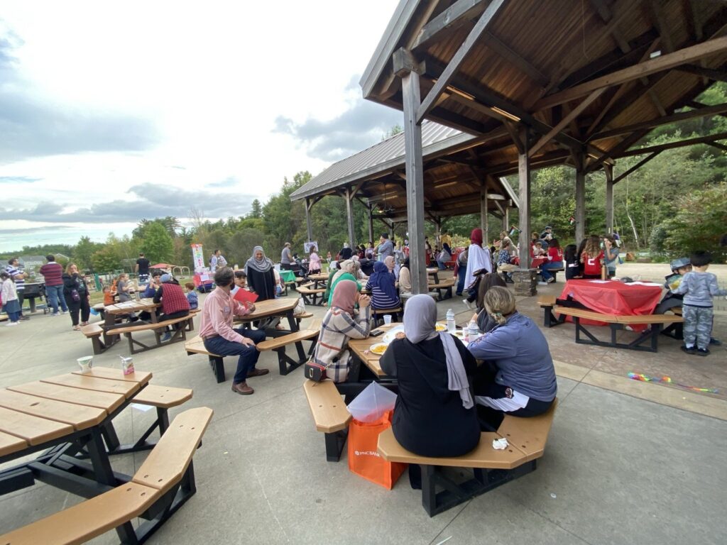 People sitting at picnic tables and walking around in the background. There are people eating, working on art projects, and chatting.