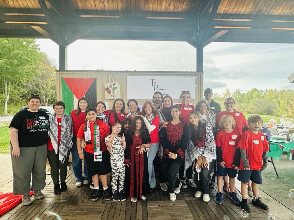 A group of children and adults pose in front of a wall that displays the Palistinian flag and a poster from The Peacemalkers Network.