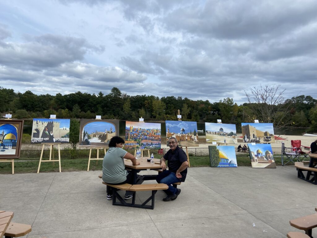 Two men sit at a picnic table. Behind them, paintings with Middle Eastern motifs (fortresses, minarets, deseert scenes) are resting on easels.