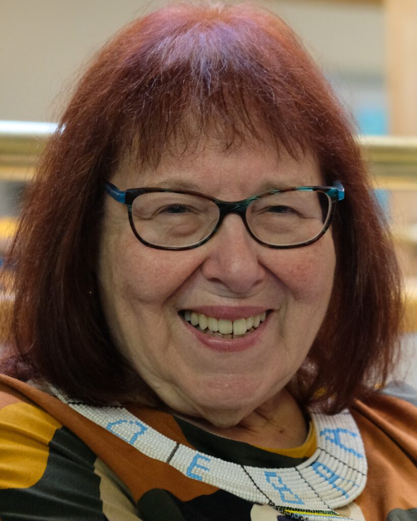 A woman with reddish hair and glasses smiles into the camera. She is wearing a white and blue beaded neckpiece.