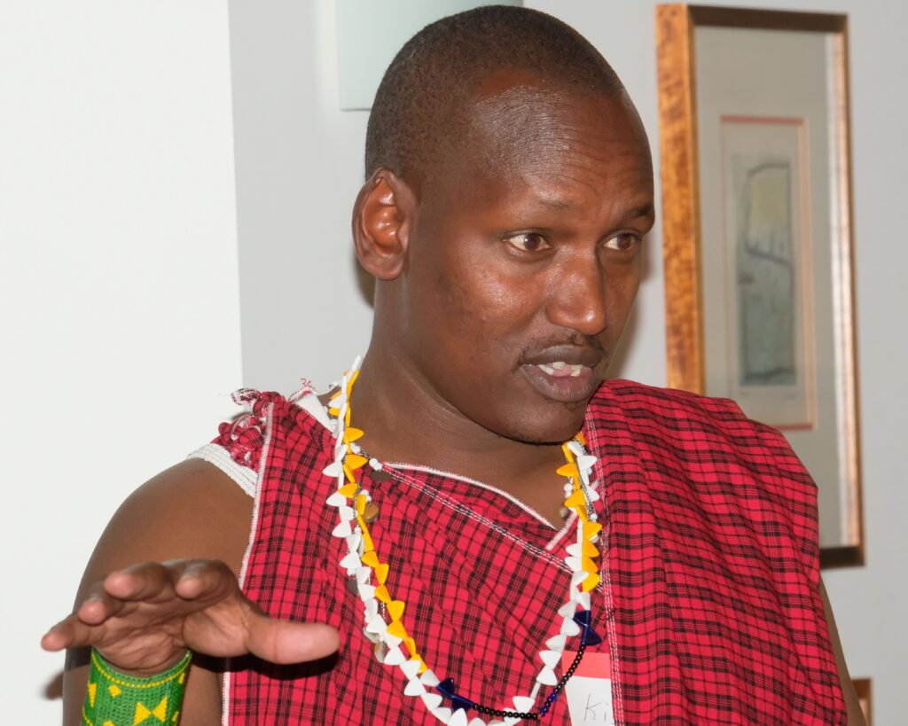 A tall black man wearing a red plaid wrap, yellow and black necklaces, and a green and yellow beaded wristband speaks to an useen audience.