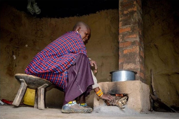 A woman wearing a plaid wrap and purple skirt sits on a low wooden stool feeds the fire in a concrete stove with a brick chimney.