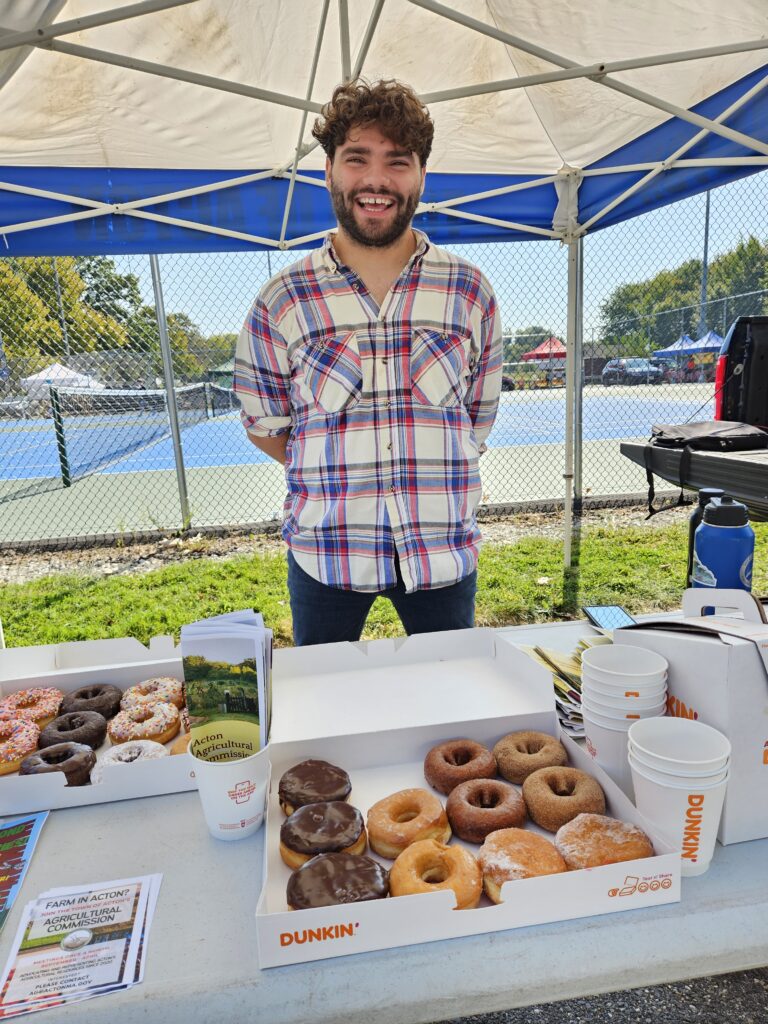 A smiling bearded man stands under a tent with boxes of donuts and Acton Agricultural Commission fliers.