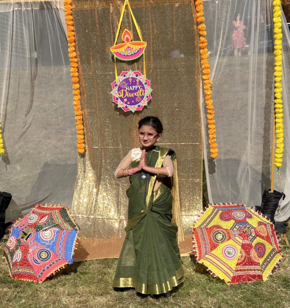 A young girl in a green and gol sari dances between two embroidered fabric parasols.