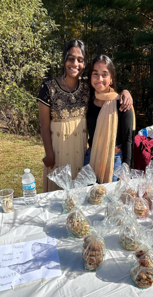 Two smiling young women, one wearing a salwar kameez stand in front of a table full of cookies for sale.