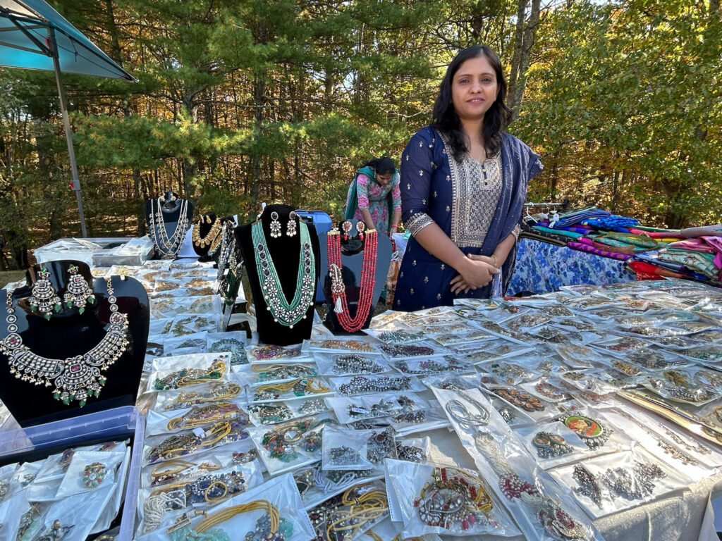 A woman in a blue salwaar kameez stands in front of a table full of jewelry sets (earrings and necklaces). A table of brighly colored sari fabric is on her right.