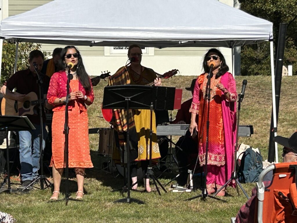 Two women wearing bright salwaar kameez sing under a tent. Musicians are visible in the background.