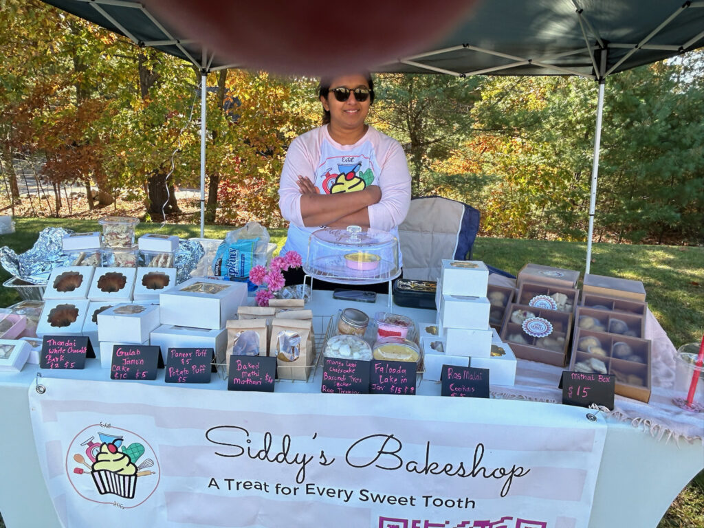 A tent with a woman in a pink t-shirt and a table full of cupcakes and other small cakes.