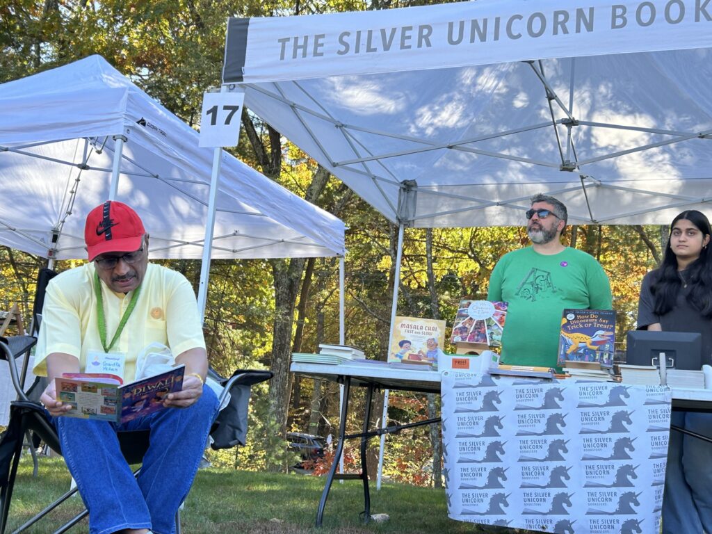 A man sits in a chair and reads a book, In the background, you can see the Silver Unicorn book tent with a couple of people behind the stacks of books.