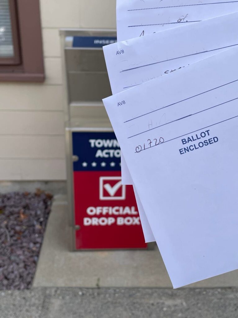 Three envelopes that say Ballot Enclosed in the foreground. The Town of Acton official ballot drop box is in the background.