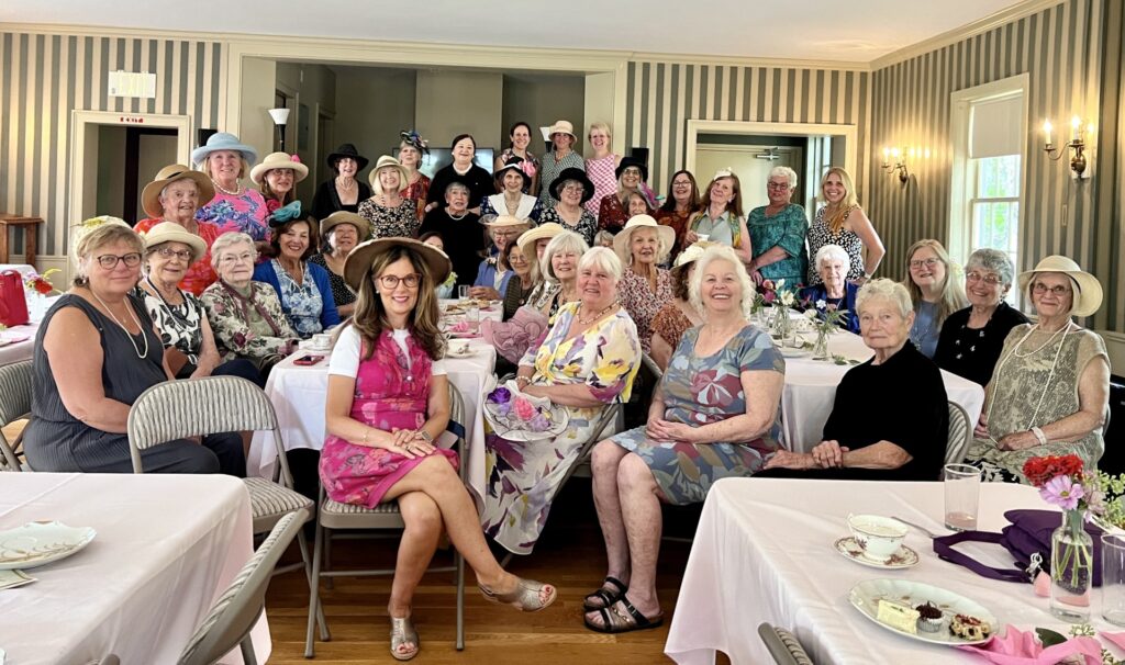 A large group of women, many wearing summer party dresses and hats, pose at one end of the Acton Womans' Club main room.