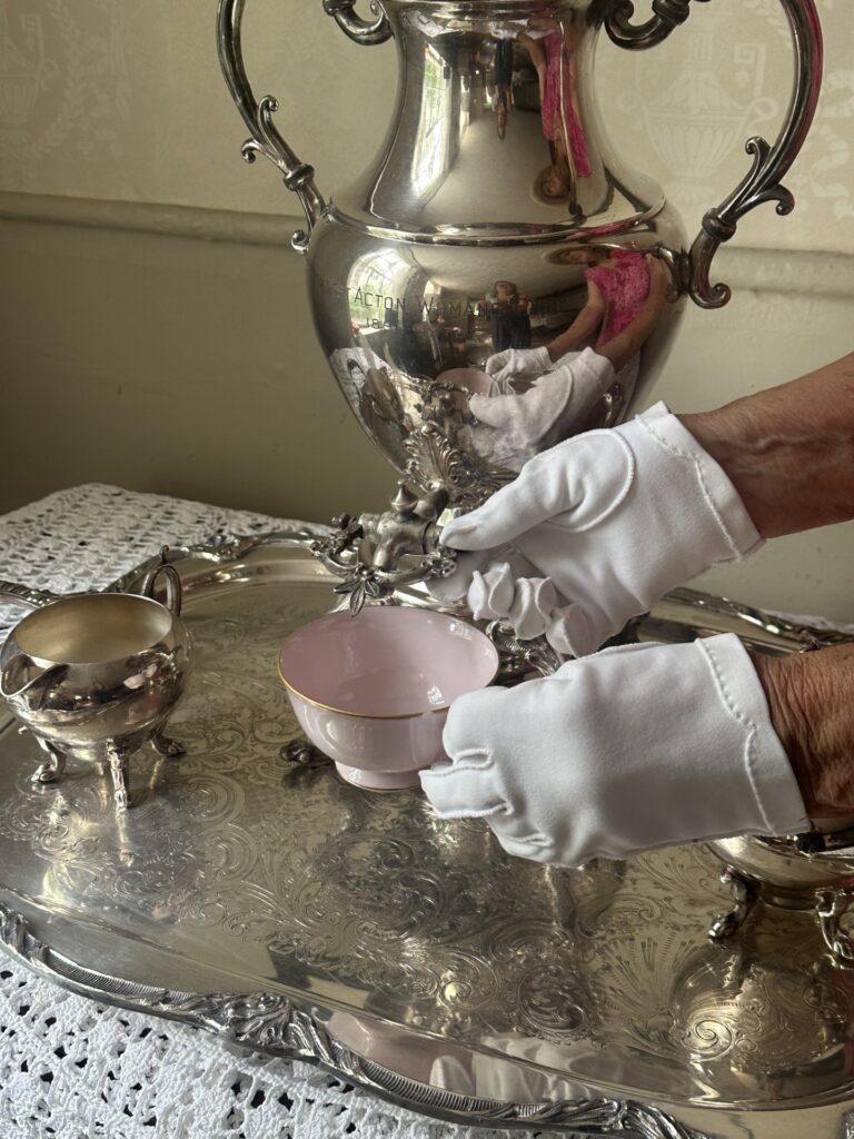 A pair of white gloved hands pours tea from a silver tea urn, which is set on a saliver tray. A silver creamer and sugar bowl are also on the tray, and the whole set is on a white lace tablecloth.