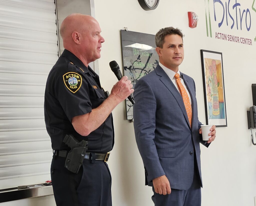 A bald man wearing an Acton Police uniform speaks into a microphone. A man in a suit and tie stands next to him (holding a cup of java).