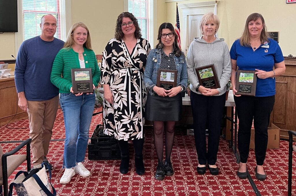 A group of people stand in Acton's Town Hall Room 204. Each person (or pair) is holding an Acton Garden Club award that includes a picture of their winning garden.