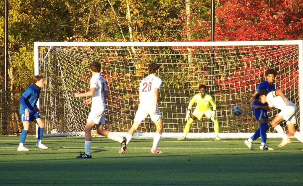 A group of high school boys play soccer in front of a net on a beautil fall day.