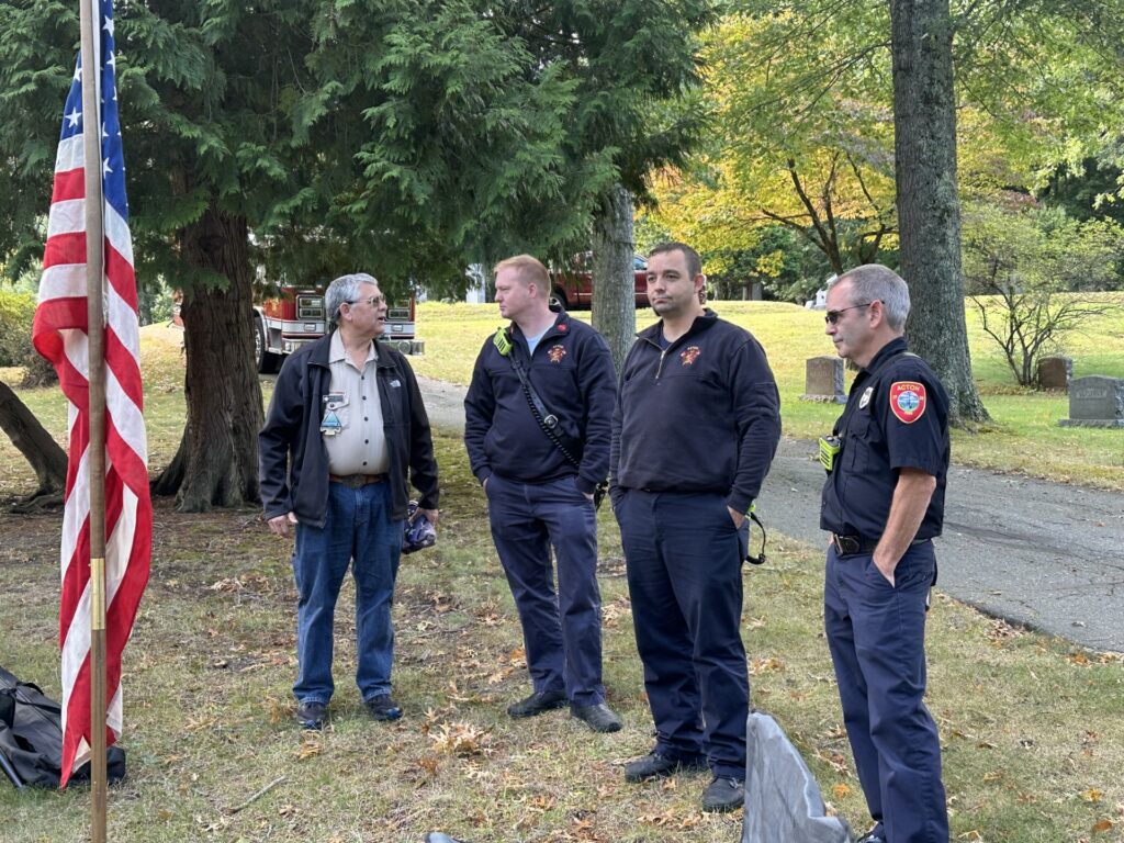 Four men in informal Fire Department clothes (shirts or jackets and jeans) stand outdoors around an American Flag.