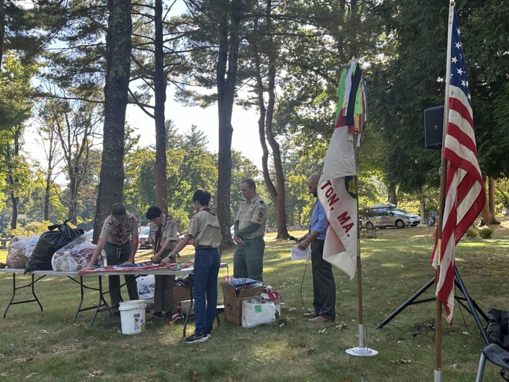 Kids in scouting uniforms work at a table outdoors. There are two flags in the foreground. 