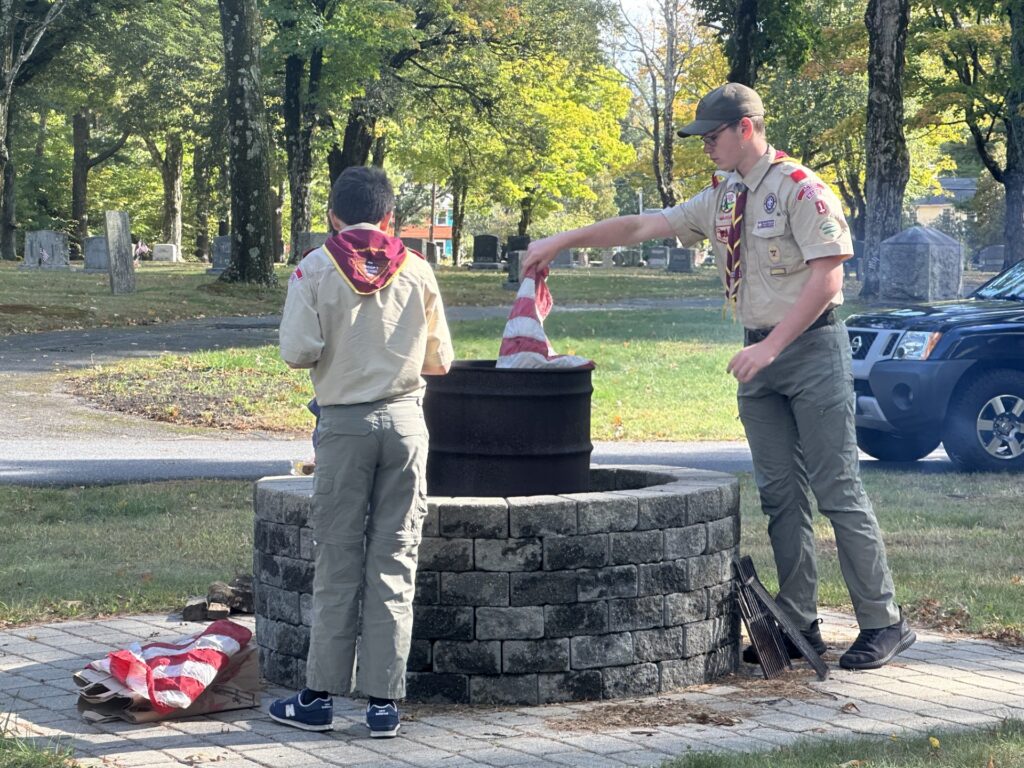 Two boys in scouting uniforms place pieces of a flag into the fire pit. The pit is a thigh-high circle of gray brick. A barrel stands in the middle (where the fire is made).