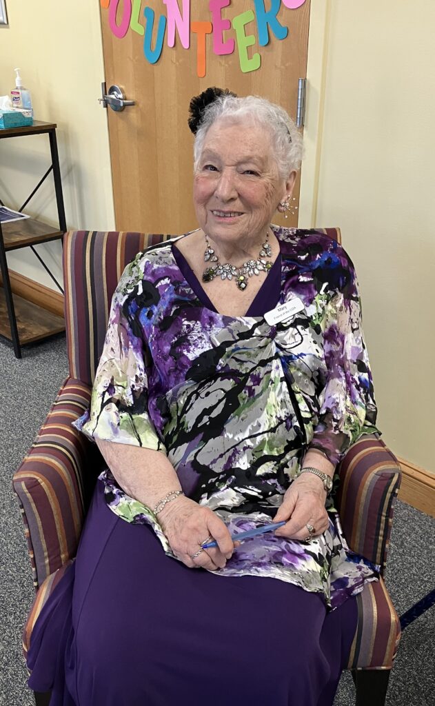 A smiling older woman with short white hair sits in a striped chair. She is wearing a purple tunic and a large (real or faux) diamond and amethyst necklace.