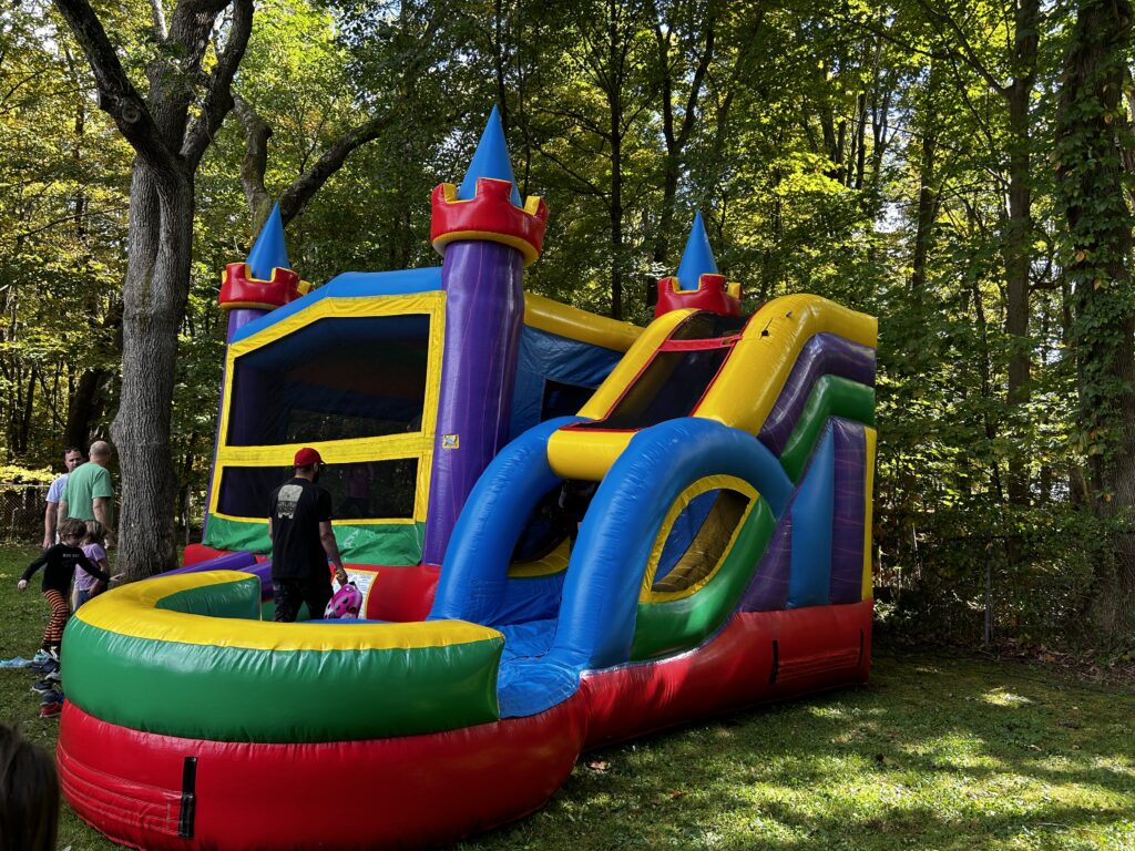 A very colorful bouncy house and slide sit under dappled shade at Gardner Field. In the background, kids line up for a turn.