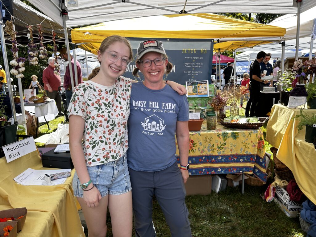 A smiling woman wearing a Daisy Hill Farm t-shirt and pigtails stands with a teenager (probably her daughter) in the Daisy Hill Farm booth. Mountain mint is for sale in the background.