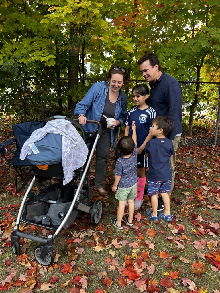 A woman on the left holds a stroller in one hand and ice cream in the other. On the right, a man and his daughter admire the contents of the stroller. Two younger boys may be more interested in the ice cream.