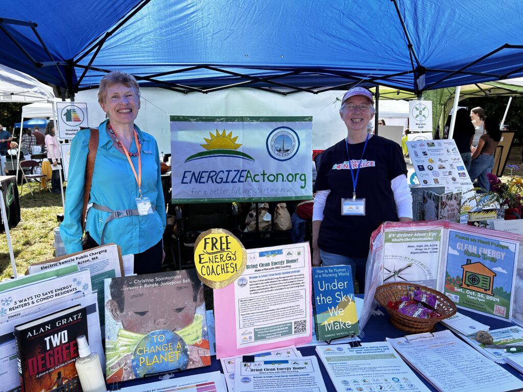 Two women stand in a booth with a large EnergizeActon banner. In the foreground is a table covered with literature and information for children and adults.