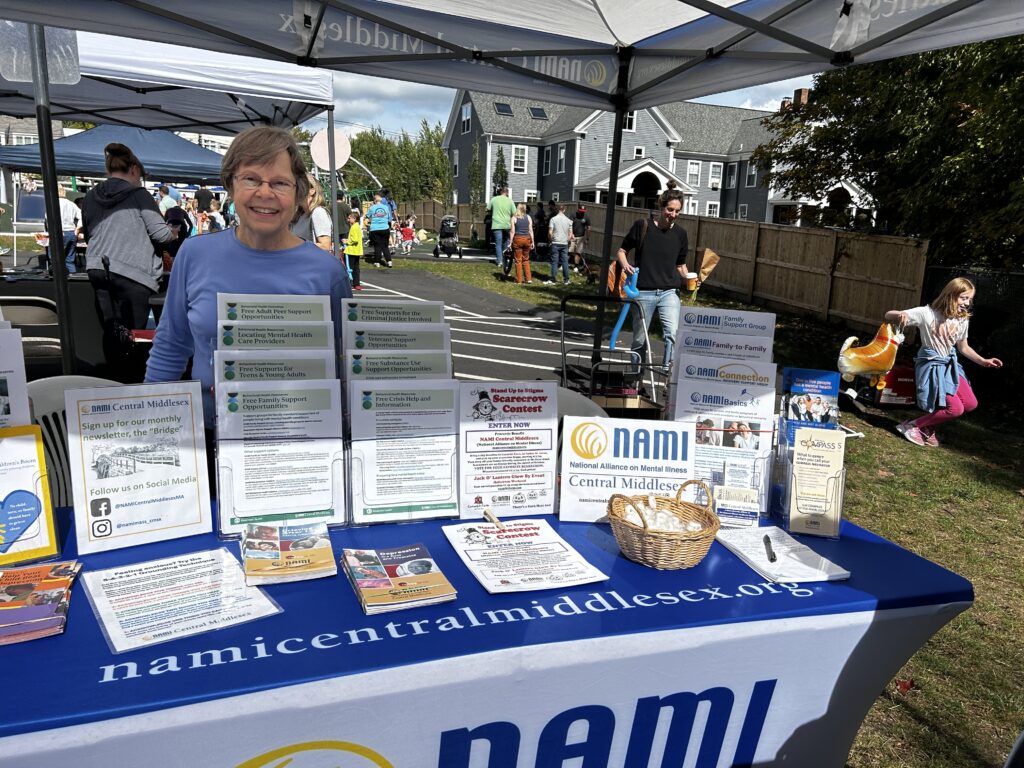 A woman stands at a table that has a NAMI (National Alliance on Mental Illness) sign. The table contains literature, a sign-up sheet and a basket wtih candy.