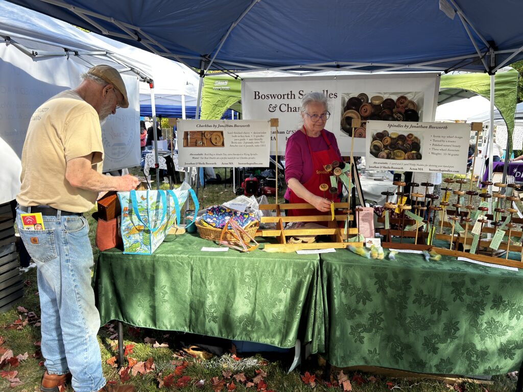 An older man and woman stand at a booth with beautiful handmade spindles (for hand-spinning wool). Each spindle is made of different wood and a work of art.