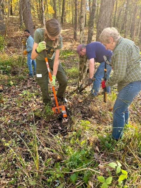 A young adult is using a large orange shovel-like tool to dig into the ground. Other people are using clippers and other tools in the background.