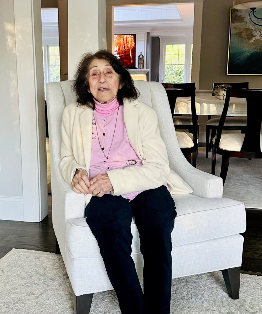 A older woman wearing a pink shirt and off-white cardigan sits in a white chair. A dining room is in the background.