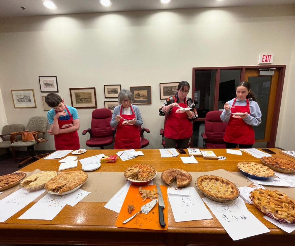 Four people wearing red aprons stand in front of a table full of pies. The boy on the right is contemplating, the other three people are eating pie. Each person has a checklist in front of them and each pie is numbered.