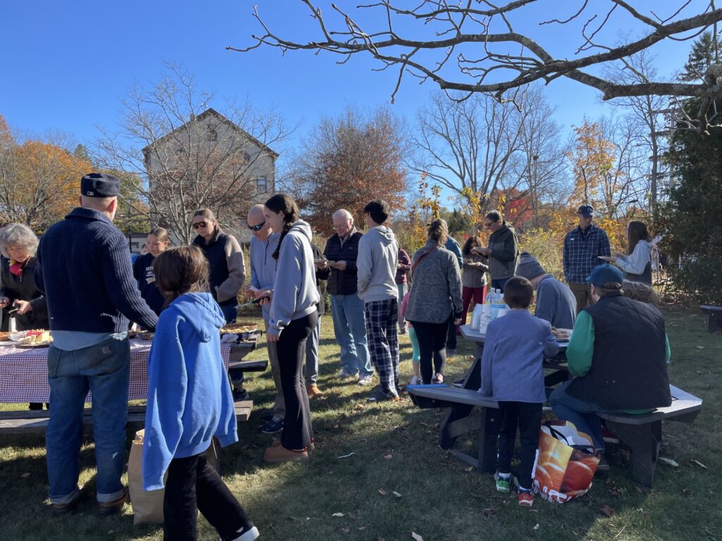 People lined up near a picnic table on a beautiful fall day. A few people are sitting at another picnic tabel enjoying the fruits of the baker's labors.