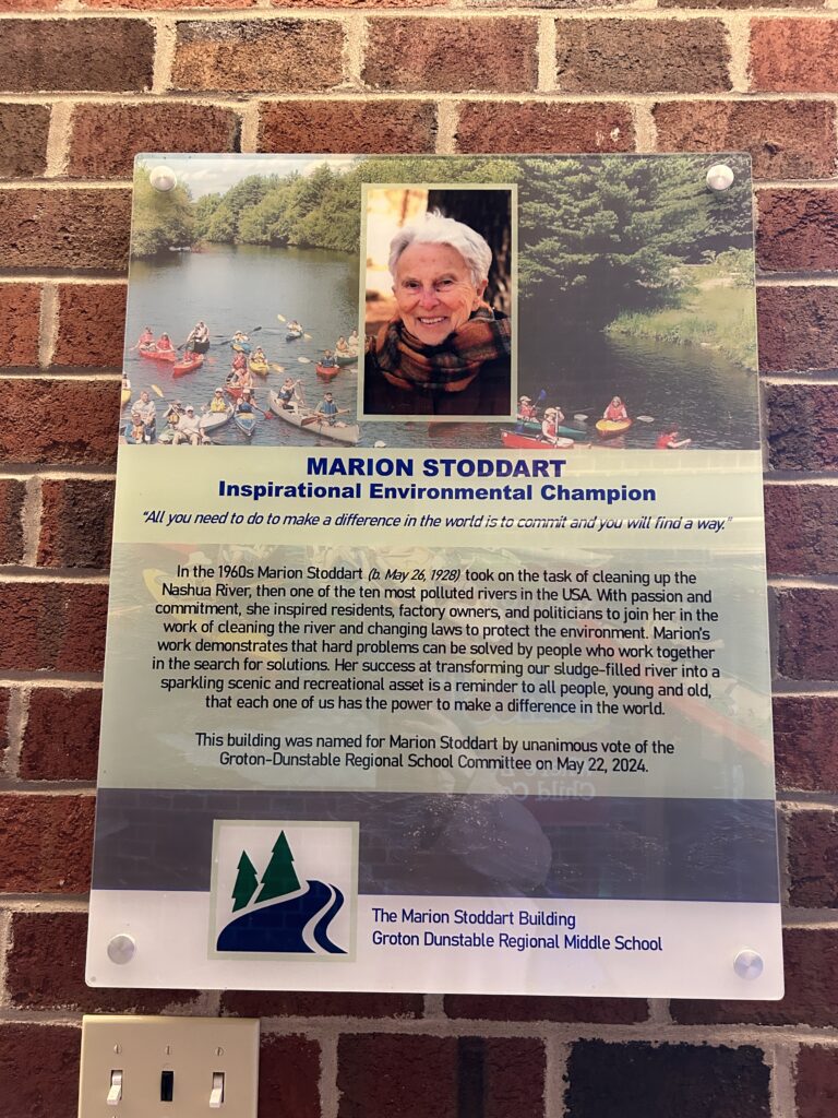 Plaque on a brick wall with a picture of Marion Stoddart over an image of kayakers on the Nashua River. The plaque also includes the following quote, "All you need to do to make a difference in the world is commit and you will find a way." A brief history of Stoddarts work and a note indicating that Marion Stoddart Building of the Groton Dunstable Regional Middle School was namedon May 22, 2024.