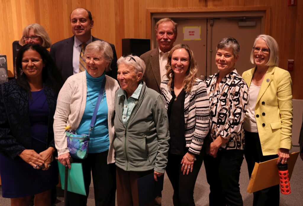 An older woman with short white hair stands with a group of other people, including State Senator Jamie Eldridge and State Rep Margaret Scarsdale.