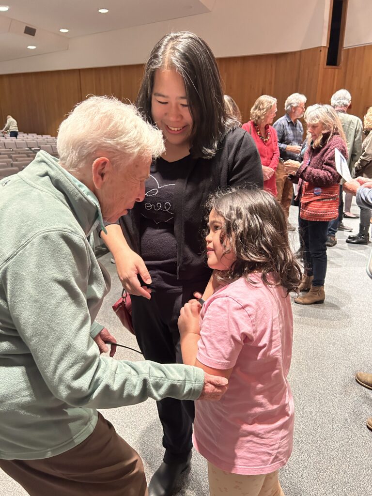 An older woman with short white hair leans down to talk to a young girl wearing a pink t-shirt. A younger woman stands behind them.