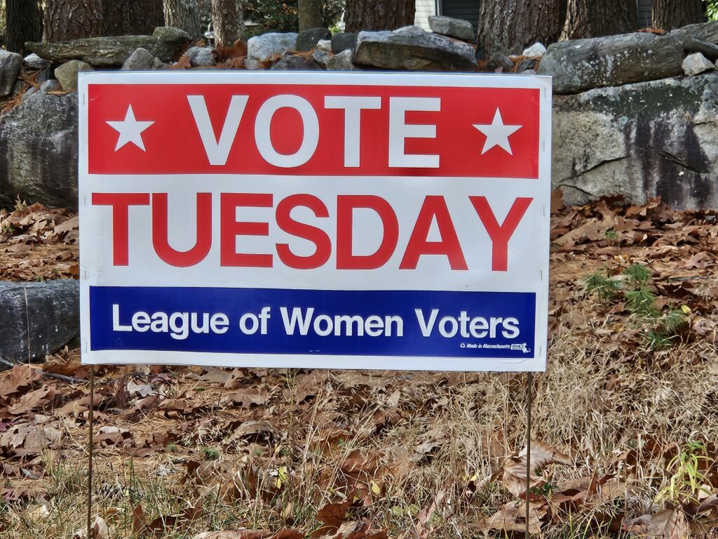 A red, white, and blue yard sign that says, " Vote Tuesday" and "League of Women Voters" planted in front of an old stone wall.