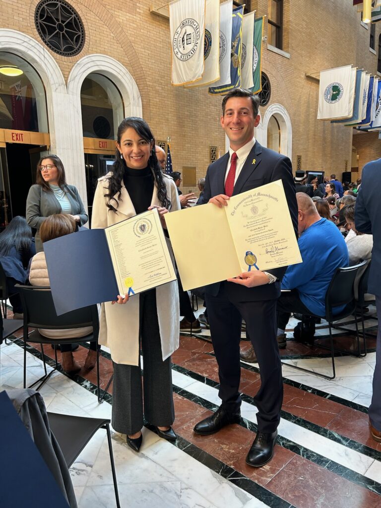 A woman with long dark hair and a man wearing a suit stand in the state house. Each of them holds a paper in a folder that has official writing and a gold seal.