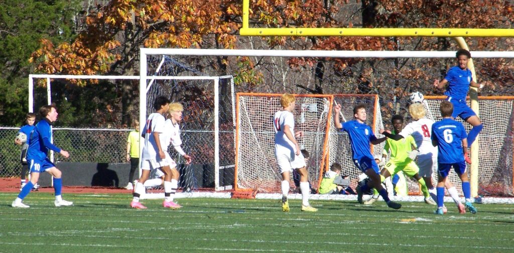 A group of young men playing soccer. One AB player is about 3 feet off the ground blocking a shot.