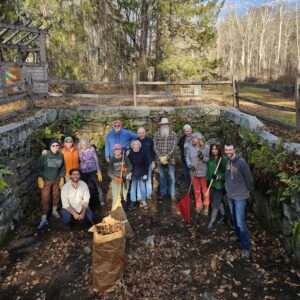 A group of people wearing work clothes and gloves stand with rakes and a large bag of debris. They are inside what was part of the mill structure. Stone walls taller than the volunteers are visible on three sides.