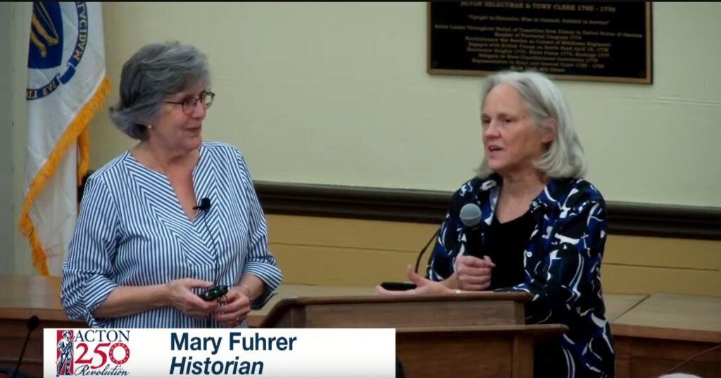 Two older women stand at a podium. The woman on the right is holding a mic.