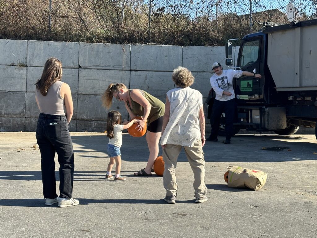 A mom holds a pumpkin for a small girl. A grandma and another person watch on the sidelines.