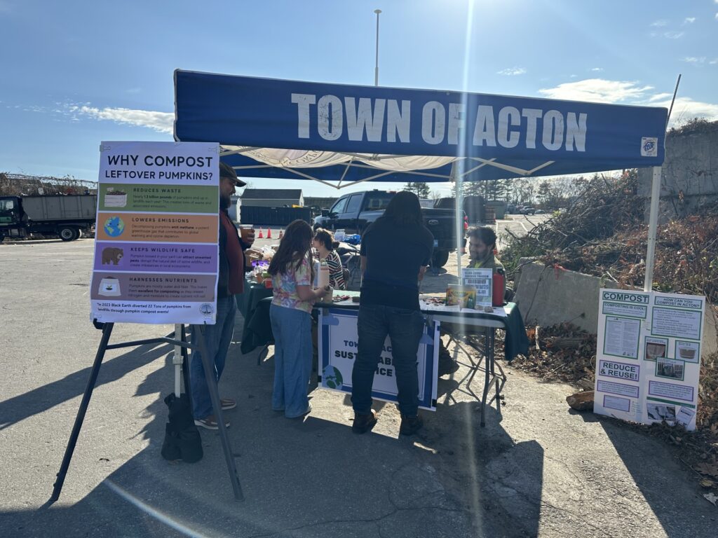 An open booth with the Town of Acton banner stands in the sun. The composting poster is on one side, and there's lots of information available. Two people stand at the booth chatting with folks.